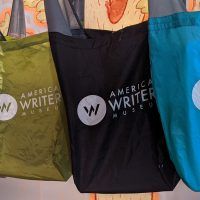 A row of hanging American Writers Museum tote bags in teal, black, green, pink, and copper