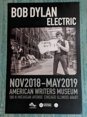 A poster from the American Writers Museum's exhibit Bob Dylan: Electric. It is black, with white text of the exhibit title above a black and white image of Dylan holding large note cards. Light grey text below the image says "NOV 2018 - MAY 2019 American Writers Museum 180 N Michigan Avenue Chicago IL 60601"