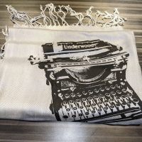 A white scarf with a black silkscreen design of an Underwood typewriter on it.