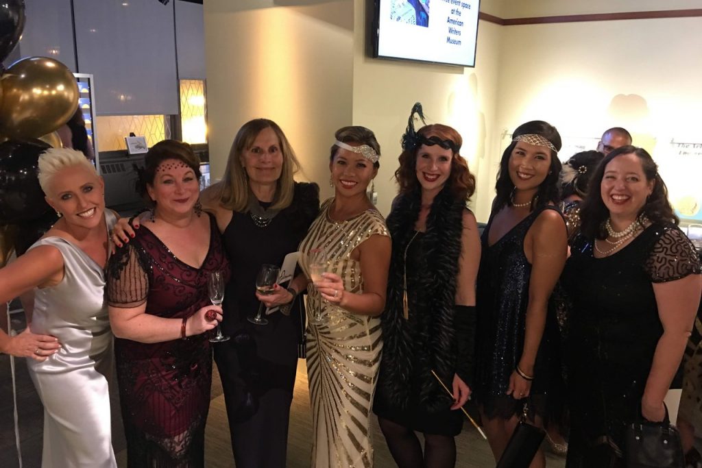 A group of 7 women dressed in 1920s inspired dresses smile at a party at the American Writers Museum