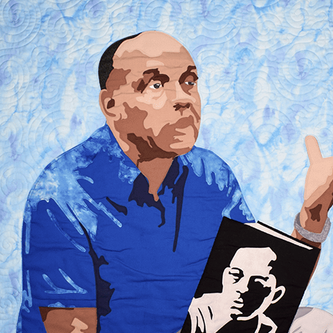 A quilt depicting Ralph Ellison seated with a cigar in one hand and his novel Invisible Man in the other. The background is light blue, with black and dark blue edges on the top and bottom.