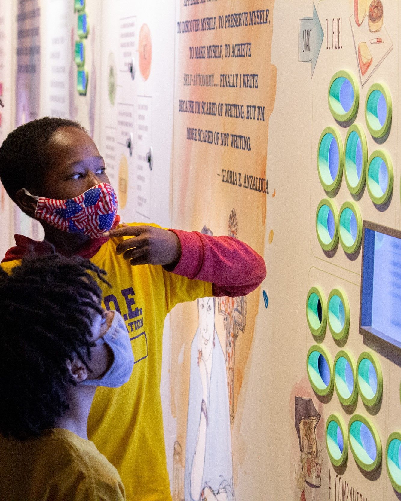 Two children on a field trip at the American Writers Museum. Both are wearing masks and looking at a wall display with multiple screens, one seated and one standing.