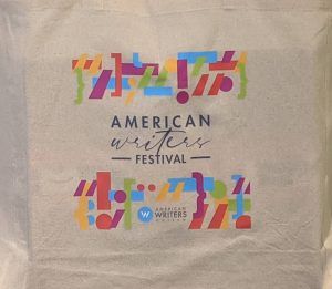 An off-white canvas tote bag with a rainbow colored punctuation design surrounding the words "American Writers Festival." The American Writers Museum logo is in the center of the lower rainbow design.